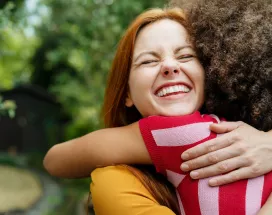 Dos chicas abrazadas, sonriendo con un fondo lleno de arboles y naturaleza.