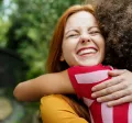 Dos chicas abrazadas, sonriendo con un fondo lleno de arboles y naturaleza.